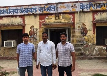 Three men stand side by side outside a faded, mural-painted building with a barred entrance and wall-mounted air conditioners.